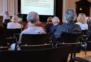 Eine Gruppe von Menschen sitzt in einem Raum und hört einem Vortrag zu. Der Vortragende steht vorne und spricht in ein Mikrofon, während eine Präsentation auf einer Leinwand zu sehen ist.