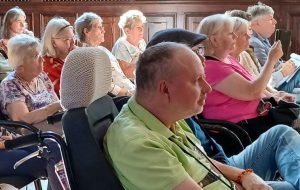 Ein Mann in einem grünen Hemd sitzt in einem Rollstuhl und nimmt an einer Tagung teil, umgeben von anderen Teilnehmern.