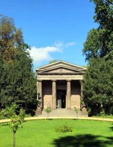 Ein klassisches Gebäude mit Säulen und Dreiecksgiebel, umgeben von üppigem Grün und Bäumen.