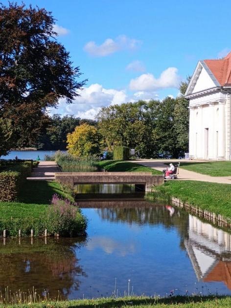 Ein ruhiger Kanal im Schlosspark Rheinsberg mit einer Person auf einer Bank und einem weißen Gebäude im Hintergrund.