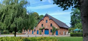 Ein malerisches Backsteingebäude mit blauen Türen und Fenstern, umgeben von üppiger Vegetation und Bäumen.