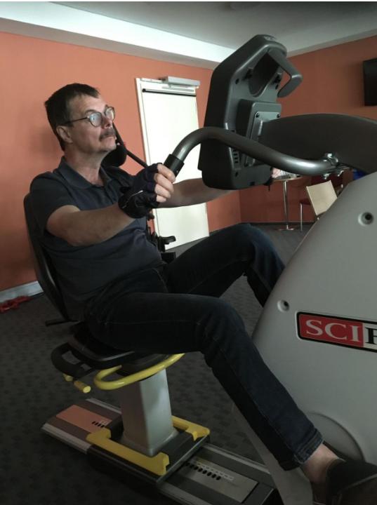 Ein Mann sitzt in einem Raum und nutzt ein spezielles Reha-Gerät, das die Armbewegungen trainiert.