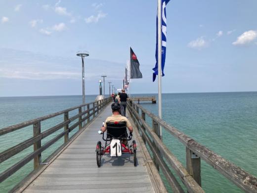 Ein Mann im Rollstuhl fährt auf einem langen Pier, der sich über das klare, türkisfarbene Wasser erstreckt. Mehrere Flaggen wehen im Wind.