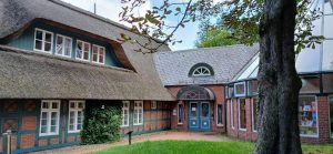 Ein malerisches Gebäude mit Reetdach und blau-weißen Fenstern, umgeben von Grünflächen und Bäumen.