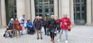 Eine Gruppe von Menschen, darunter einige im Rollstuhl, steht vor dem Eingang des Humboldtforums in Berlin. Die Gruppe wirkt fröhlich und gesellig.