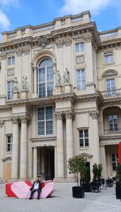 Ein Mann sitzt auf einer modernen Bank vor dem imposanten Gebäude des Humboldt-Forums, das mit klassischen Säulen und Skulpturen verziert ist.