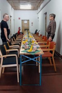 Zwei Männer stehen in einem Raum mit einem langen Tisch, der mit gelben Dekorationen und Snacks gedeckt ist. Der Raum ist mit Stühlen und Tischen ausgestattet.