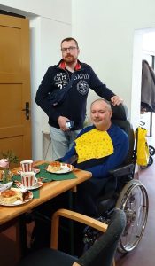 Zwei Männer, einer im Rollstuhl, essen gemeinsam in einem Raum. Der eine trägt eine gelbe Serviette am Hals.