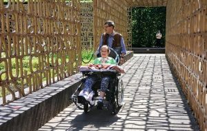 Eine Person im Rollstuhl wird durch einen Garten mit Bambus-Architektur geschoben. Die Sonne scheint und wirft Schatten auf den Weg.
