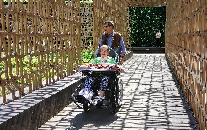 Eine Person im Rollstuhl wird durch einen Garten mit Bambus-Architektur geschoben. Die Sonne scheint und wirft Schatten auf den Weg.