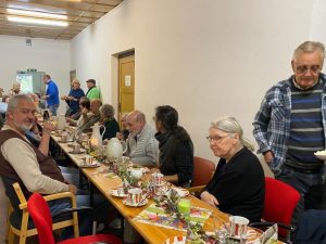 Eine Gruppe von Menschen sitzt an einem langen Tisch und genießt ein gemeinsames Essen. Der Tisch ist mit Tellern, Tassen, einer Kanne und Dekorationen geschmückt.