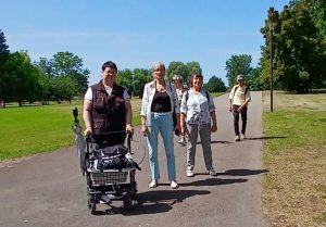 Eine Gruppe von Menschen geht auf einem Weg in einem Park spazieren. Eine Person schiebt einen Rollator.